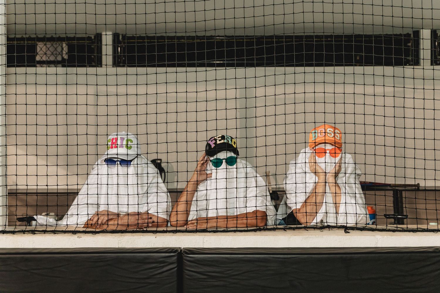 Figures in ghost costumes sit behind baseball field netting at night.