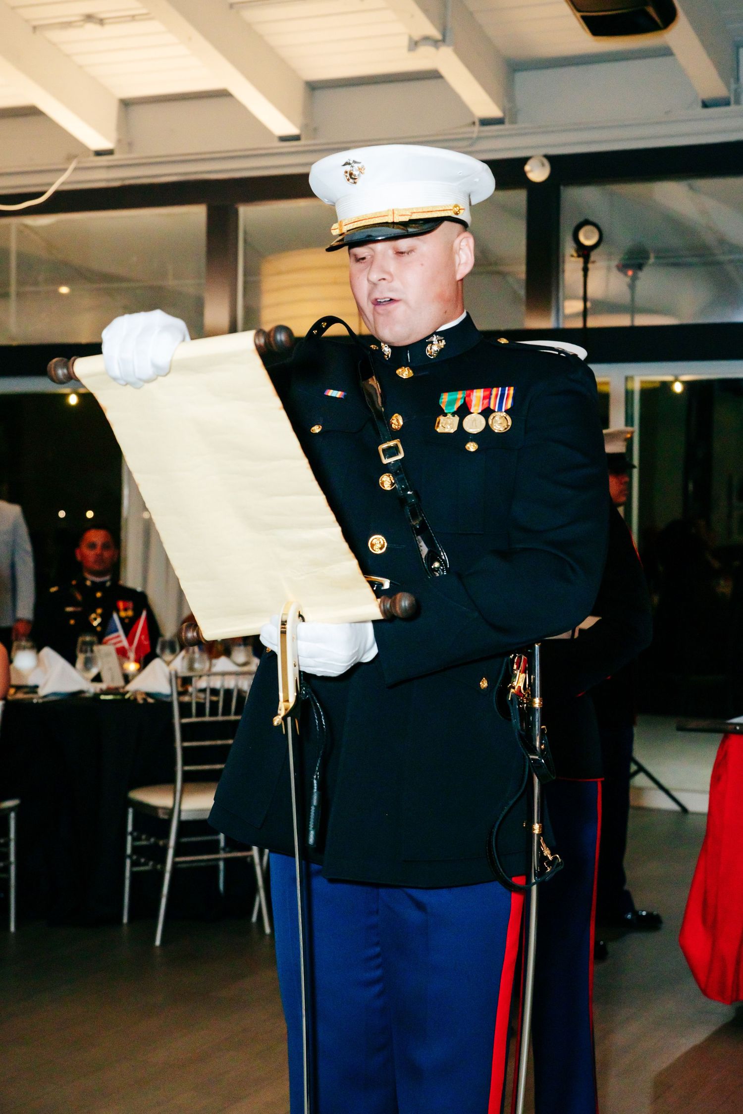 Marine in dress uniform reads from ceremonial scroll during formal military event.