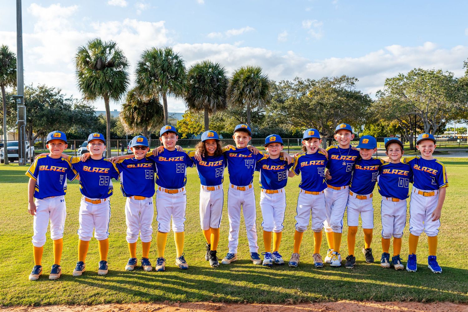 Baseball team in bright blue jerseys and white pants stands in line on a sunny field with palm trees in the background.