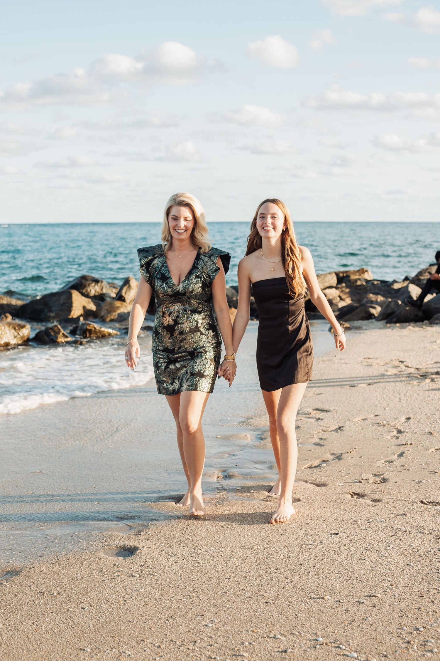Two people strolling barefoot along a sandy beach with rocky coastline and blue ocean views.