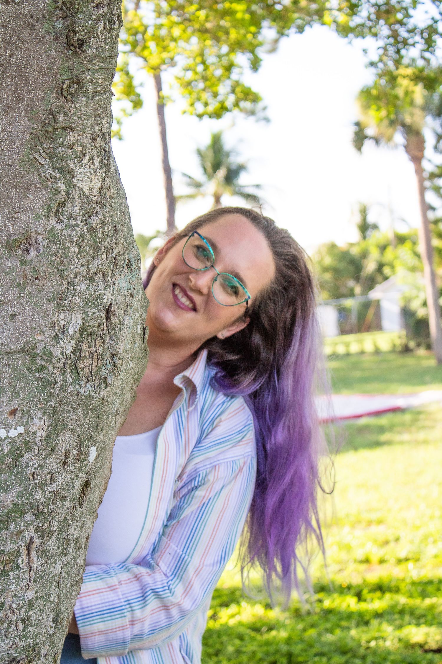 Cheerful artist in pastel striped shirt posing by tree trunk in sunny garden.