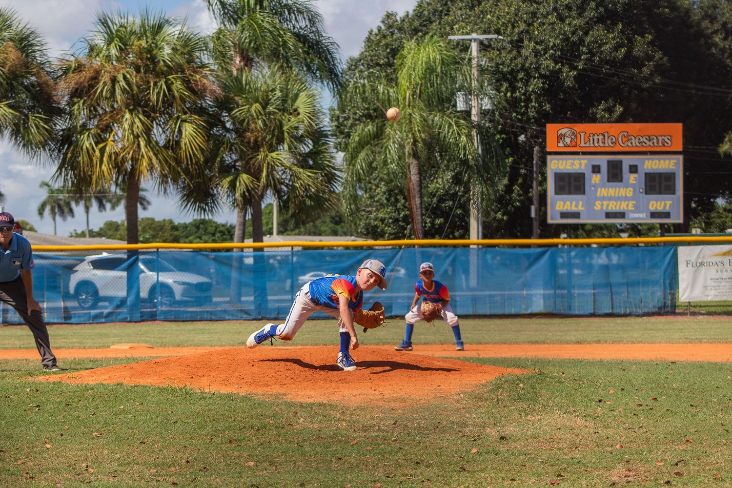 Youth baseball players in action on a sunny field with palm trees and scoreboard in the background.