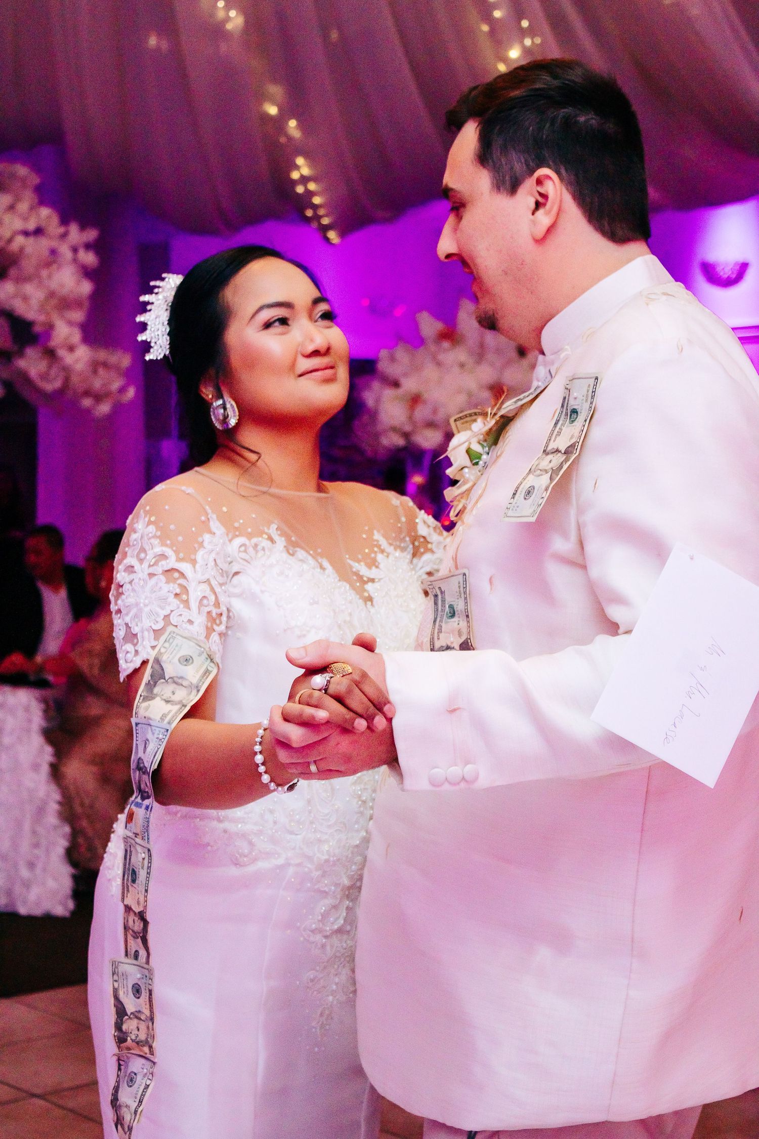 Close-up of newlyweds dancing together against pink-lit floral backdrop.