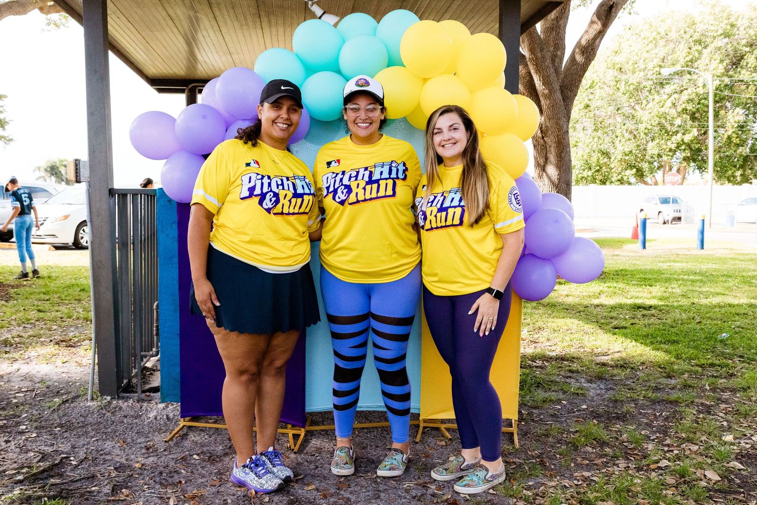 Three supporters in yellow Long Beach shirts pose together in front of colorful rainbow balloons at an outdoor event.