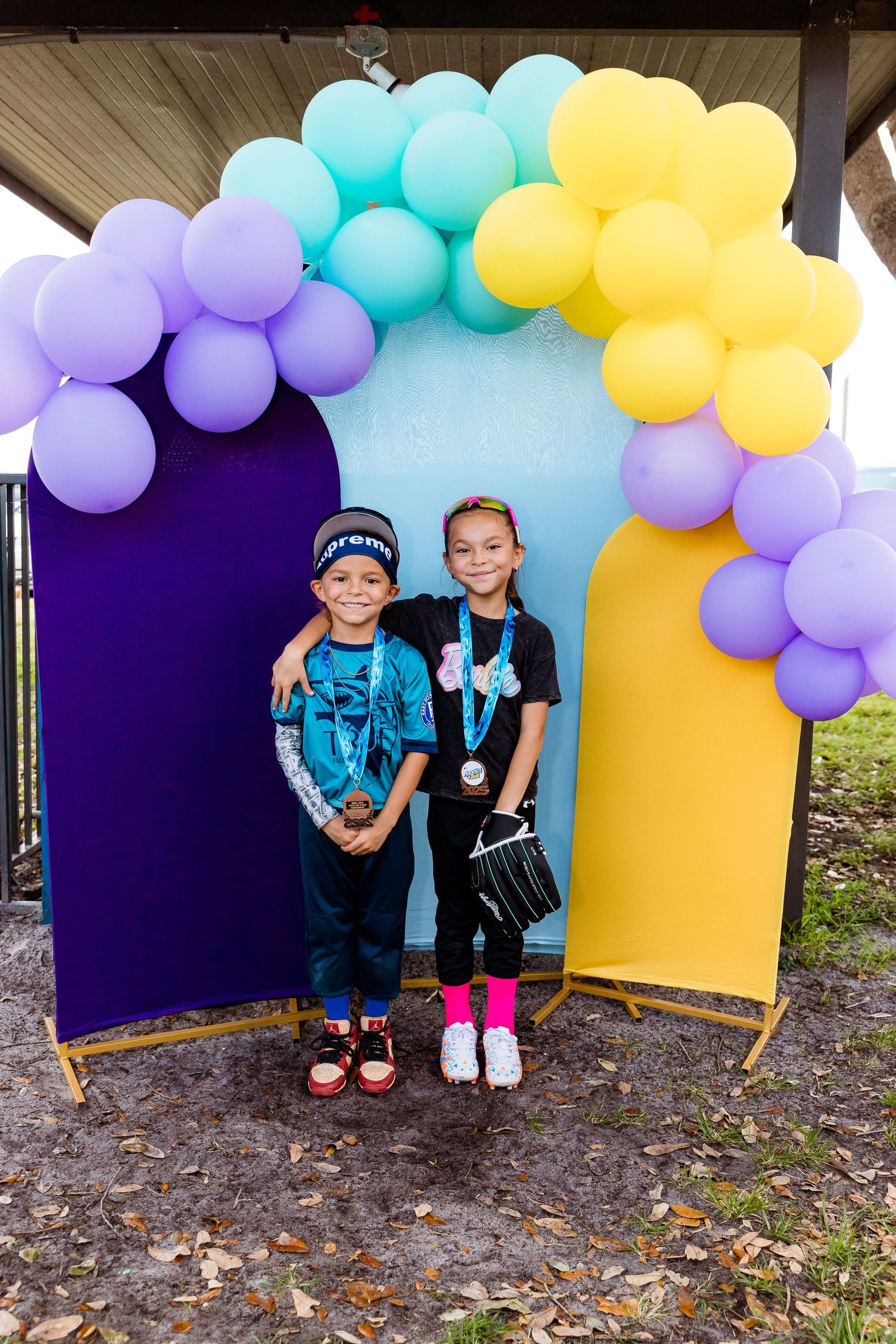 Colorful balloon arch with purple, turquoise and yellow balloons decorates a photo backdrop display at an outdoor event.