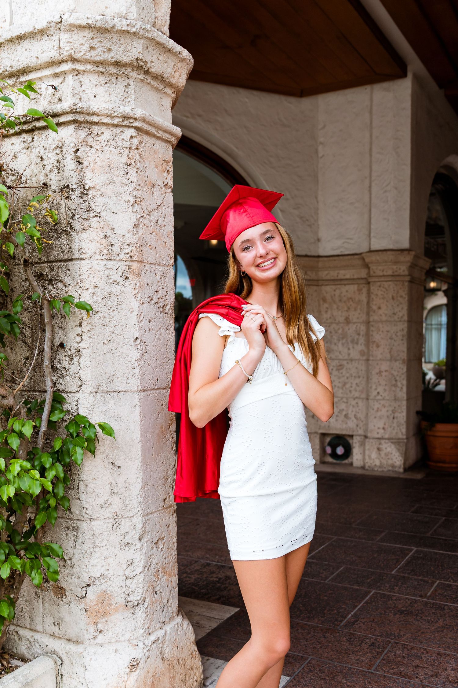 Graduate in white dress and red cap poses by stone pillar with ivy during graduation photo session outdoors.