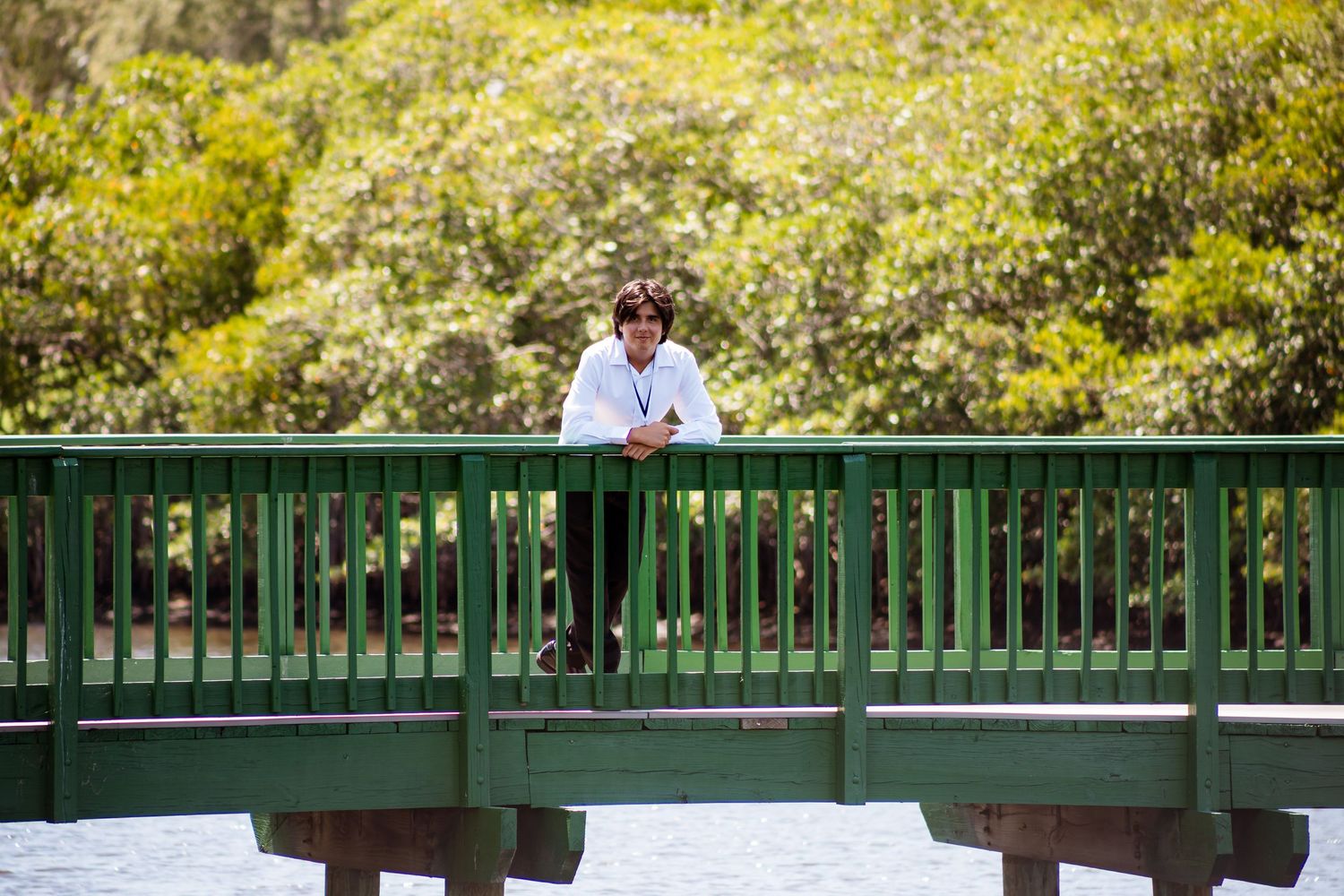 A person wearing white stands on a green painted footbridge surrounded by lush yellow-green foliage.