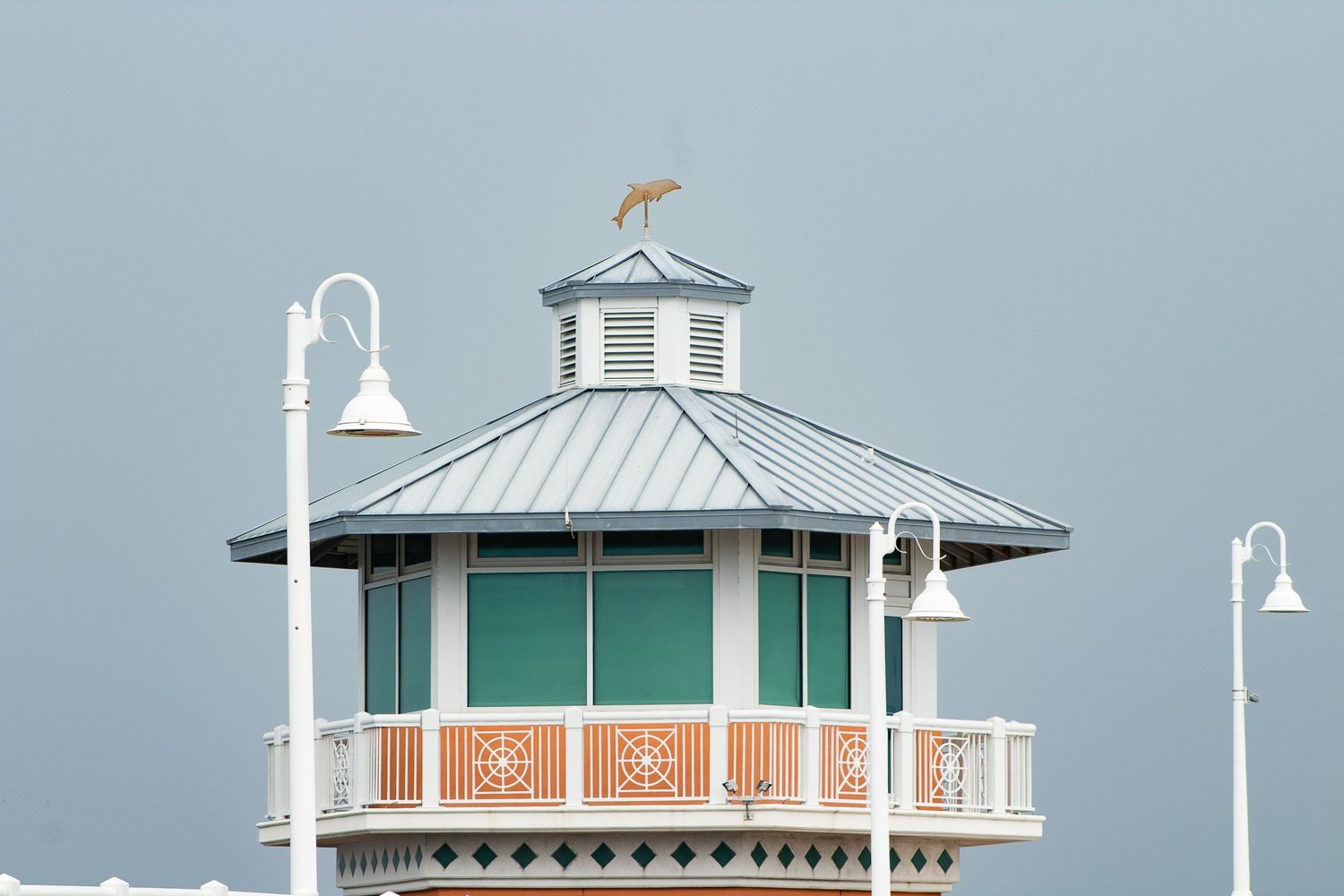 A weathered coastal gazebo with teal windows and white lamp posts against a gray sky features a seabird perched on top.