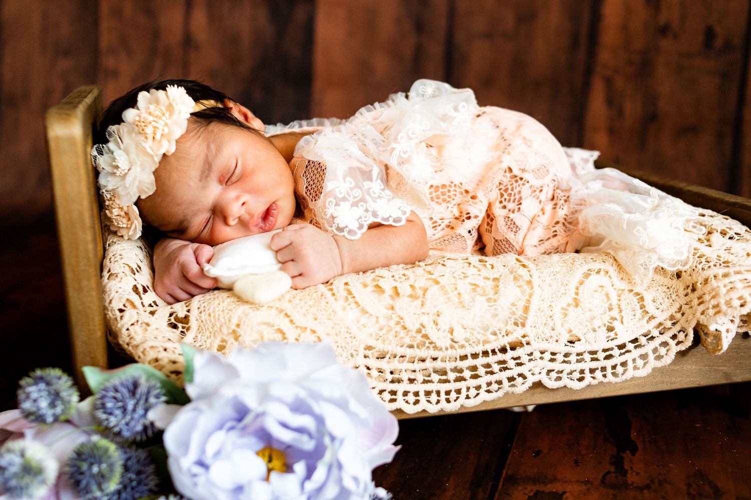 Sleeping newborn baby in lace dress and floral headband on crochet blanket in wooden bed with flowers.
