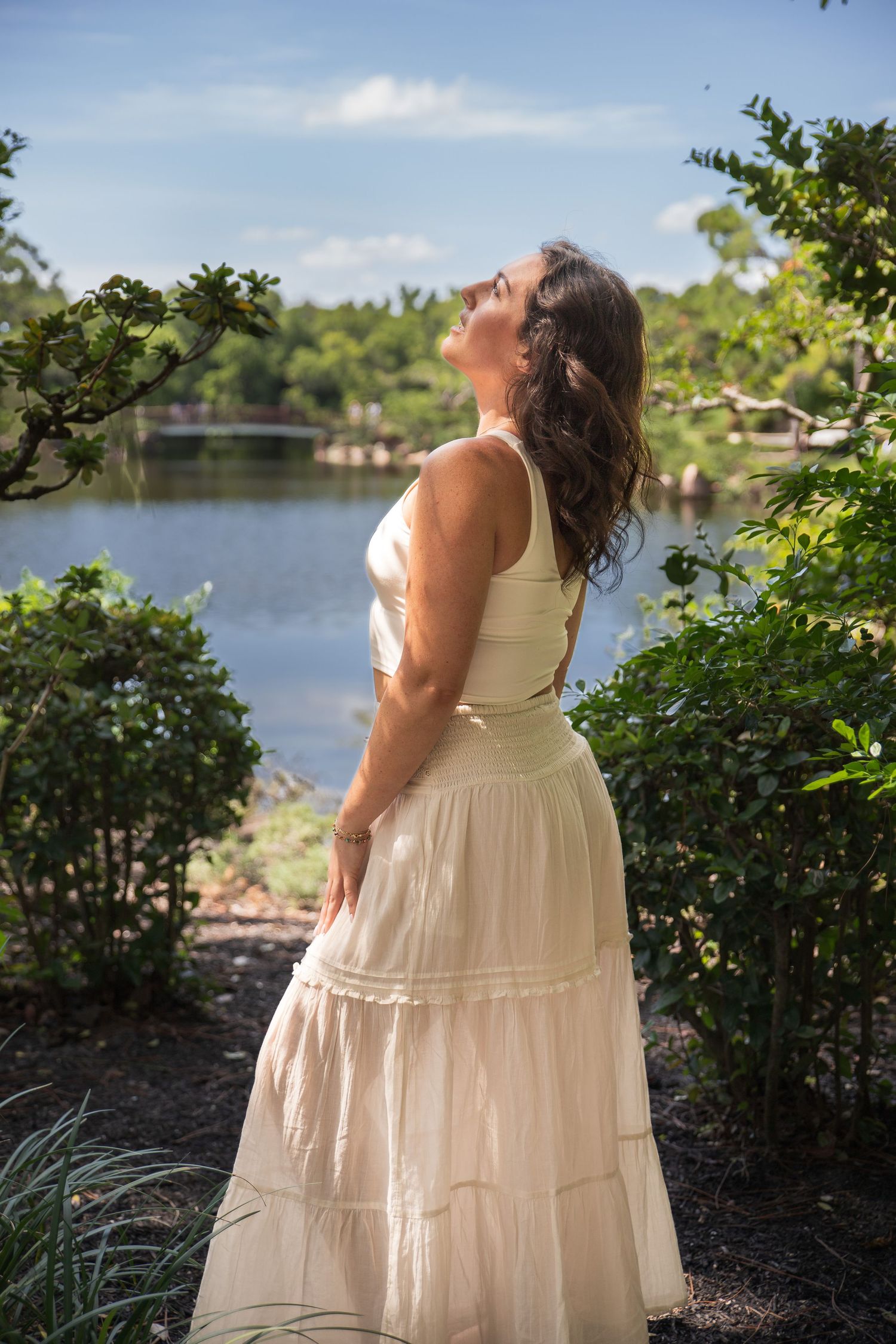 A person in a flowing white dress stands beside a lake on a sunny day among greenery and bushes.