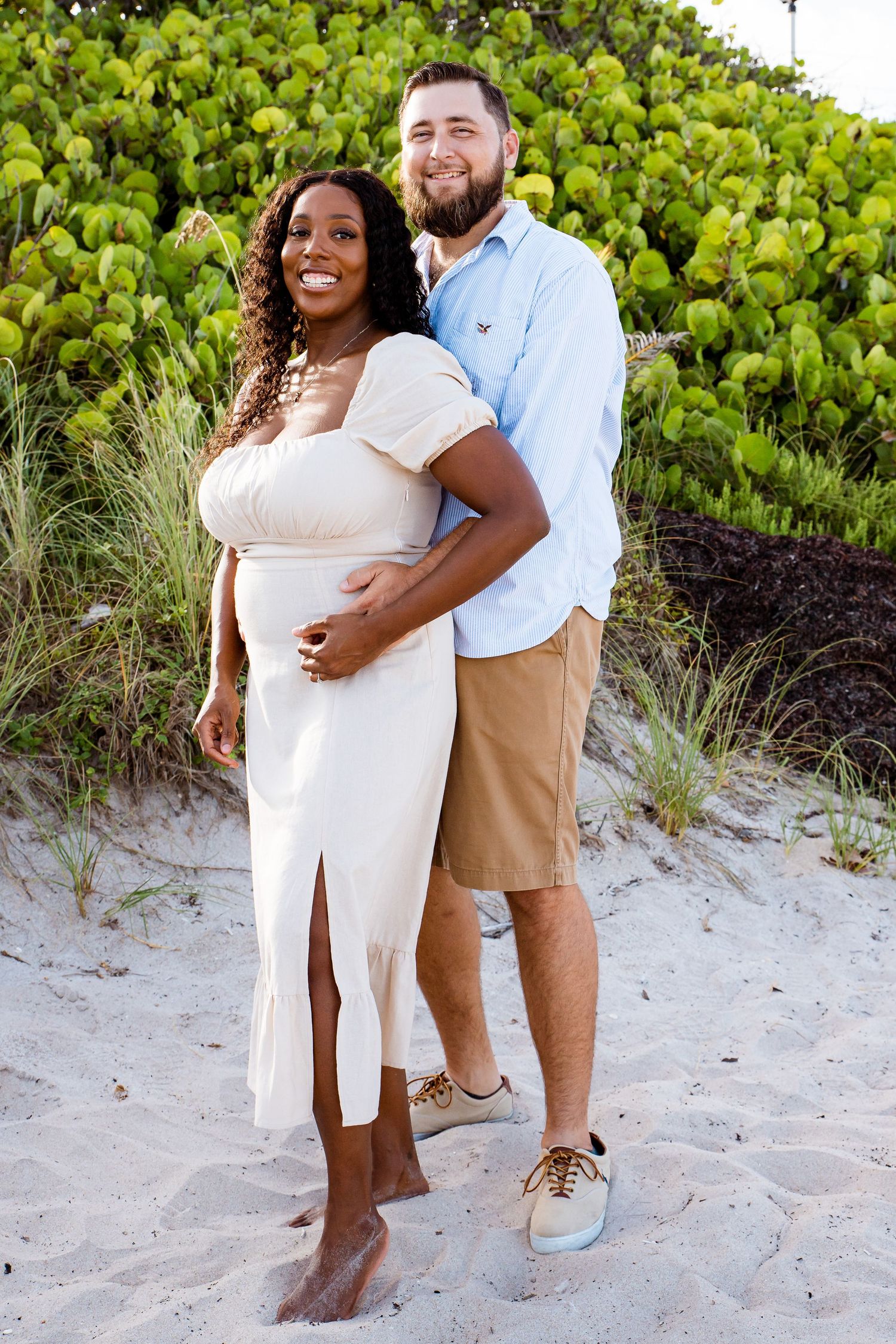 Couple poses together on a sandy beach with green foliage in the background during a sunny day.
