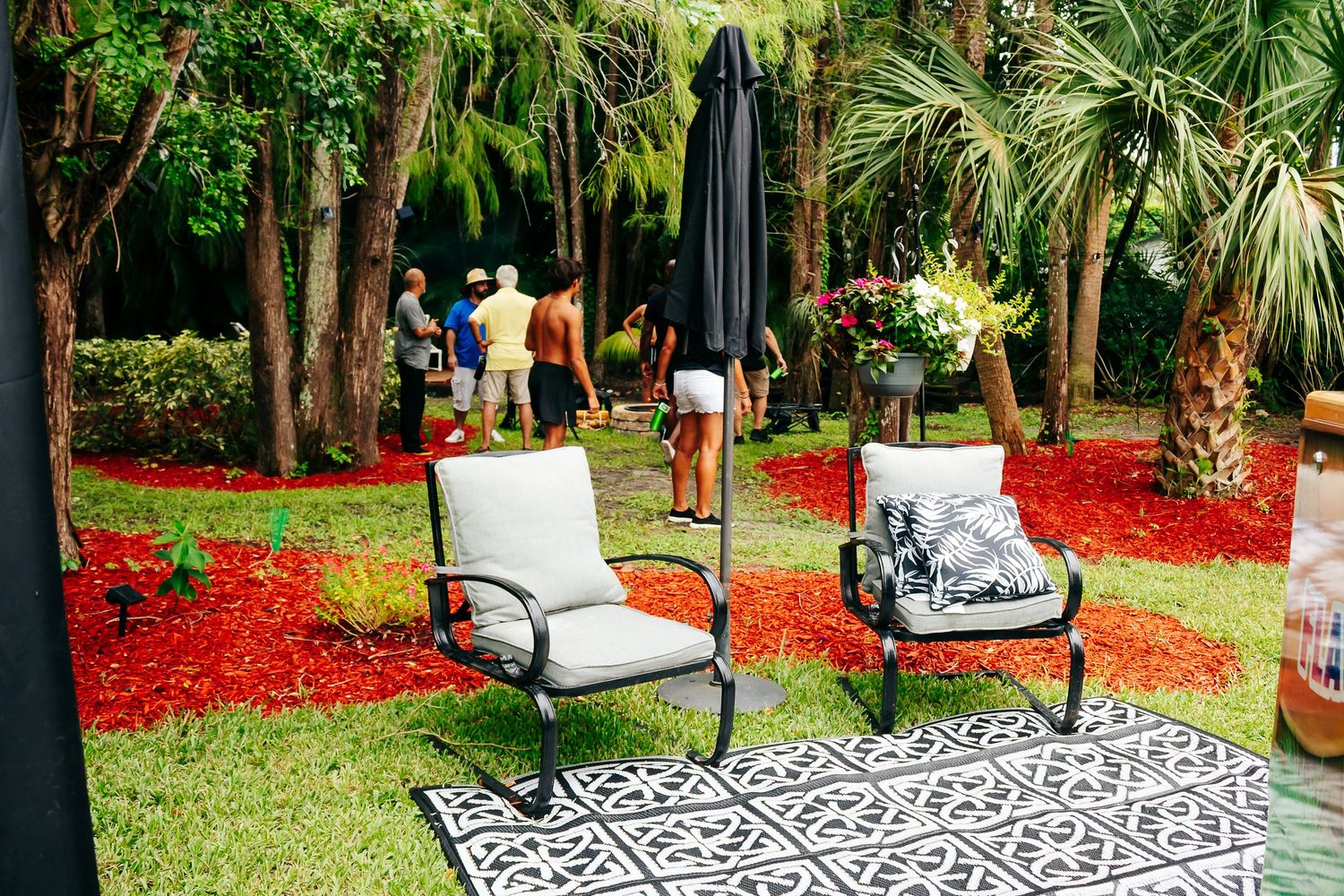 Patio seating area with geometric rug and chairs set up on red mulch with palm trees.