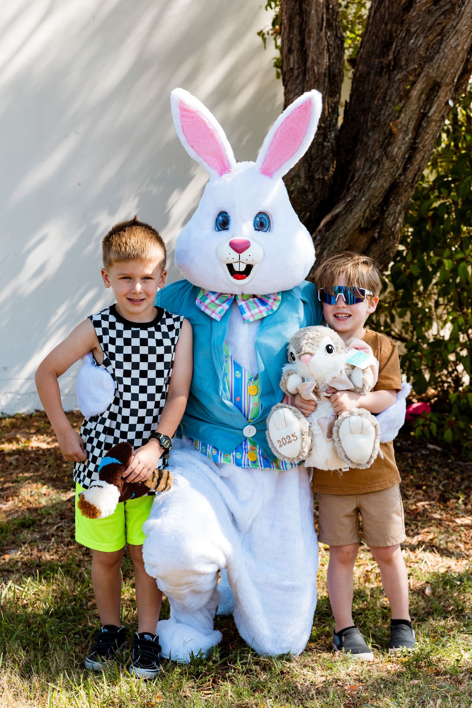 Life-sized Easter Bunny character in blue vest poses with children holding baskets and plush toys at springtime event.
