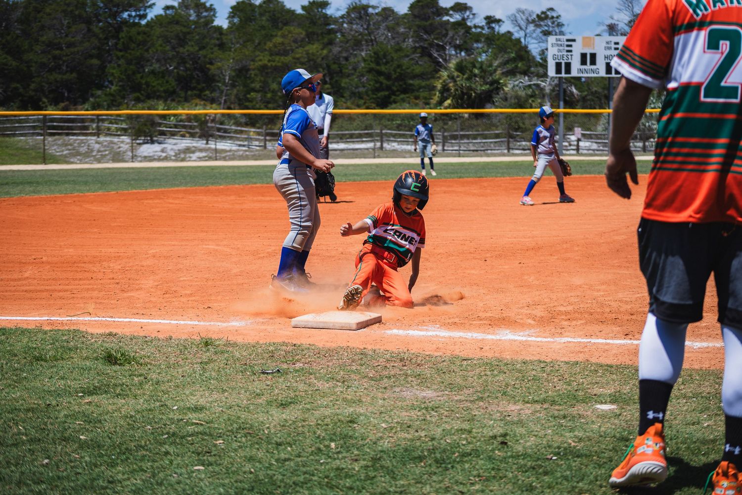 A baseball player slides into base while a fielder attempts to make the play on a dirt diamond field.