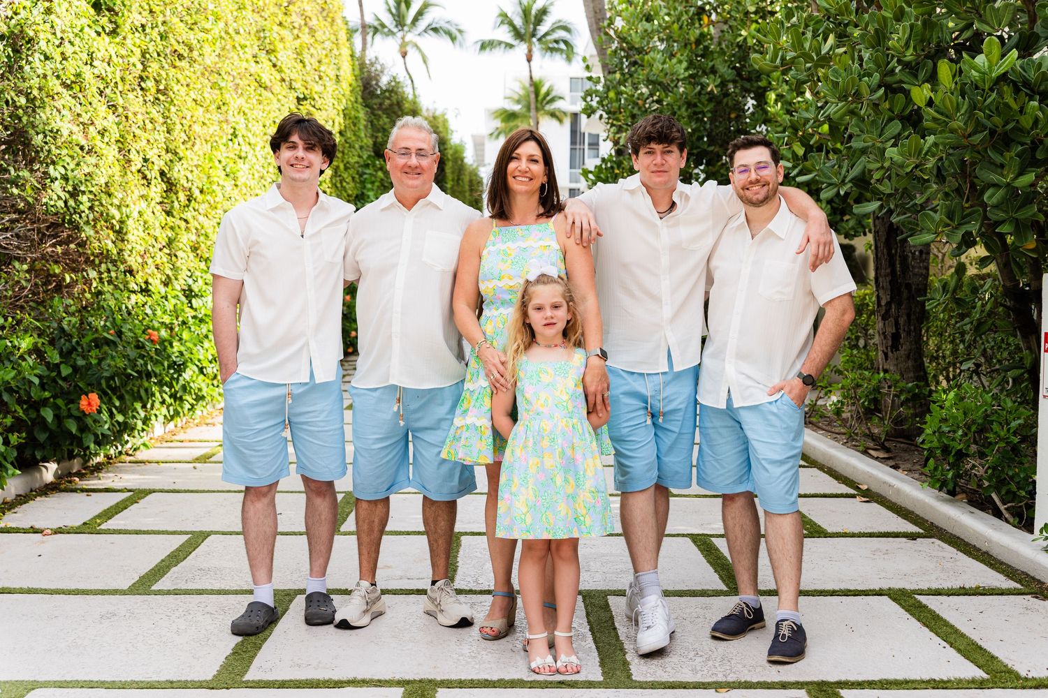 A group of people in coordinating light blue and white outfits pose together on a palm-lined pathway.