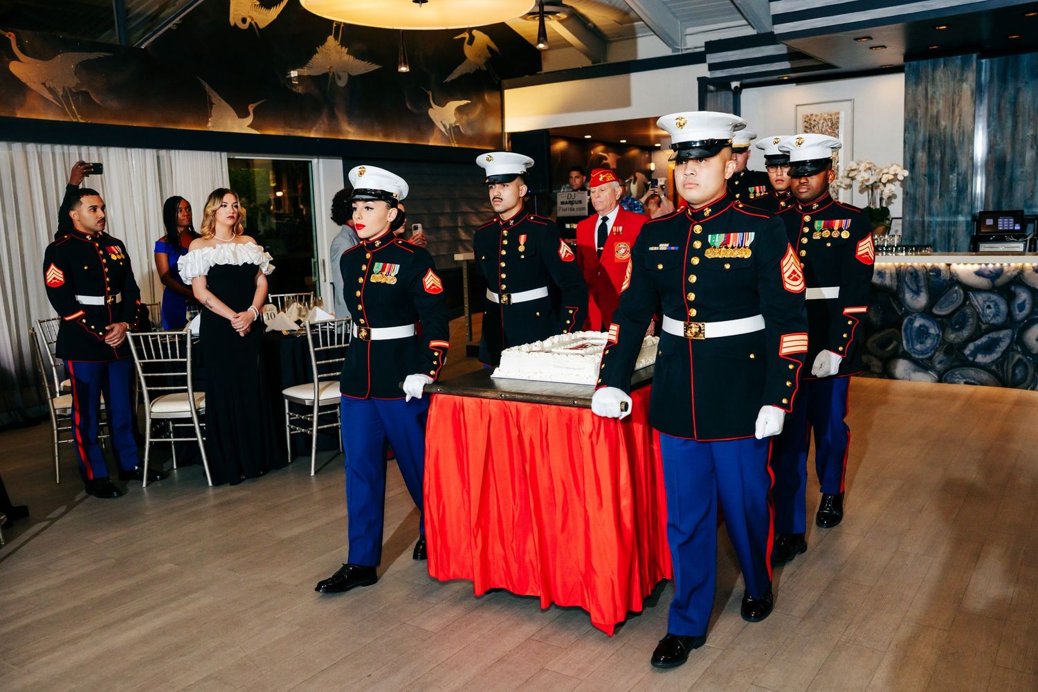 Military personnel in dress uniforms participate in a formal ceremonial event with red tablecloths and decorative settings.