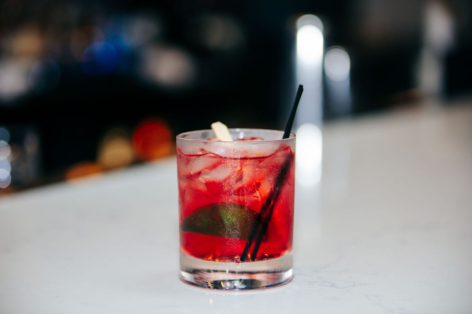 A red cocktail with ice and mint leaves served in a rocks glass with a black straw on a white surface.