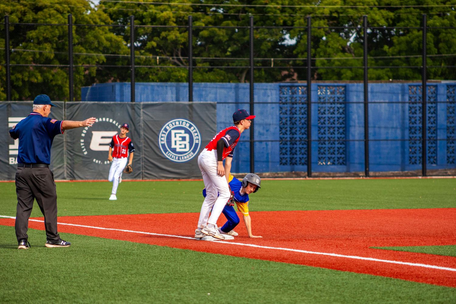 Baseball players at base during a youth game, with umpire signaling on a sunny day at a blue-fenced field.
