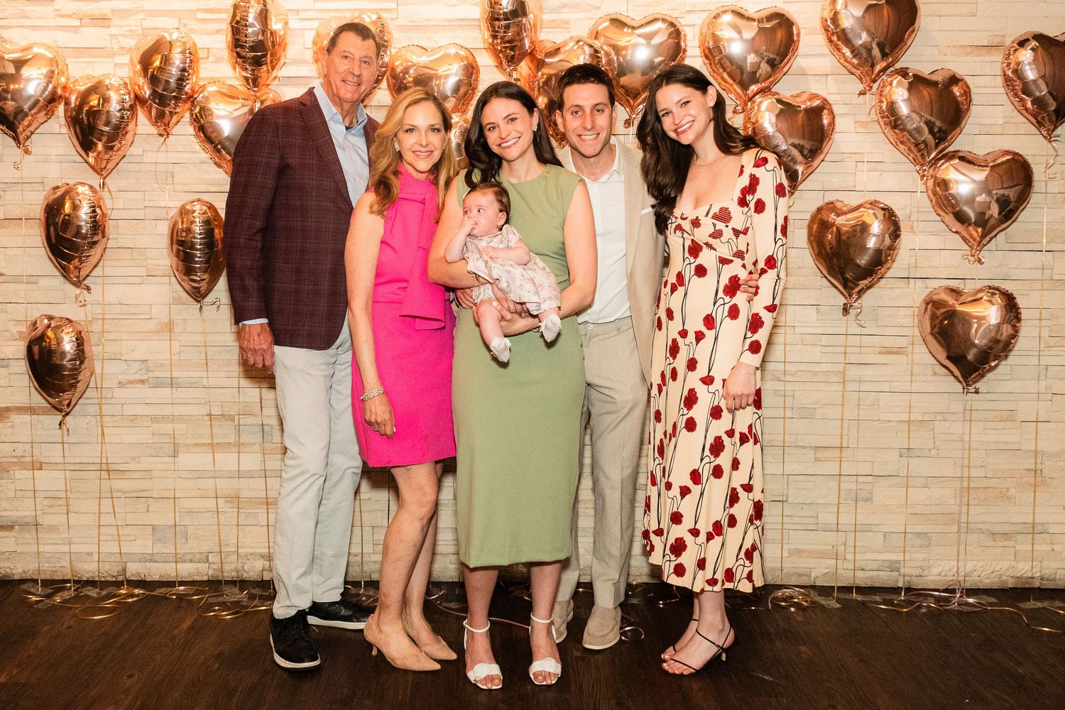Group photo at celebration with rose gold heart balloons against brick wall backdrop.