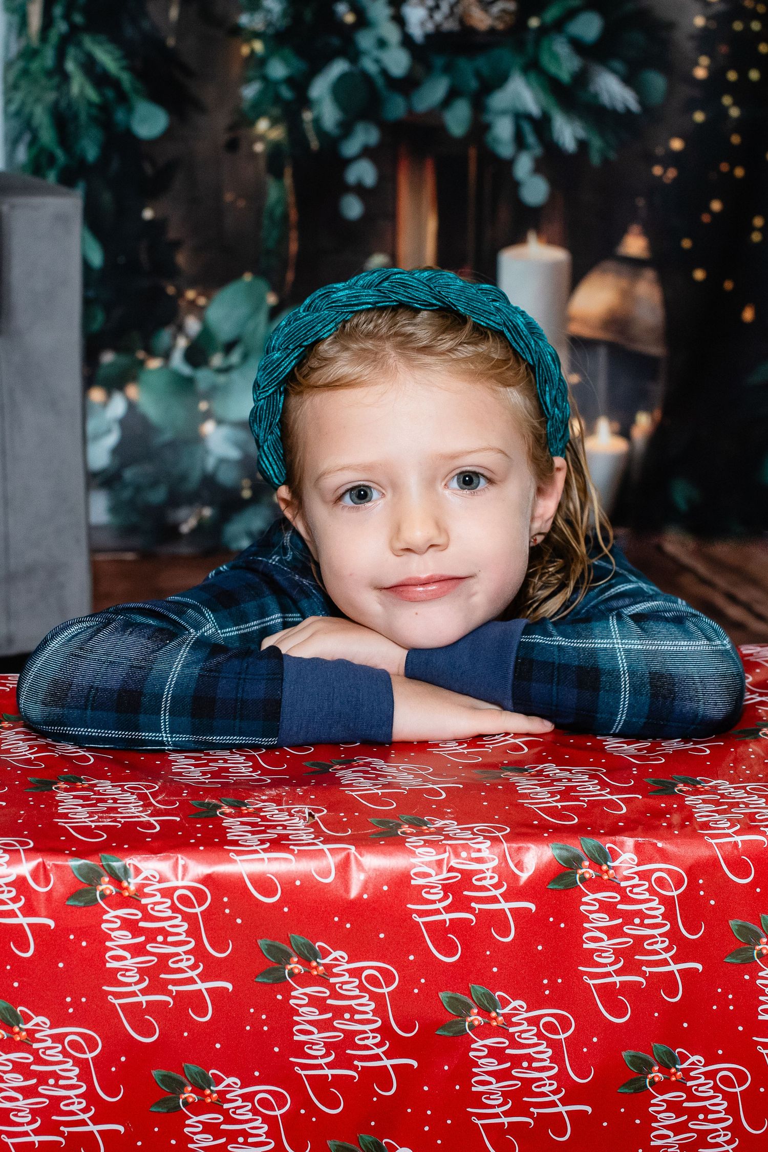 A child in plaid pajamas and teal headband rests on a Christmas-patterned surface.