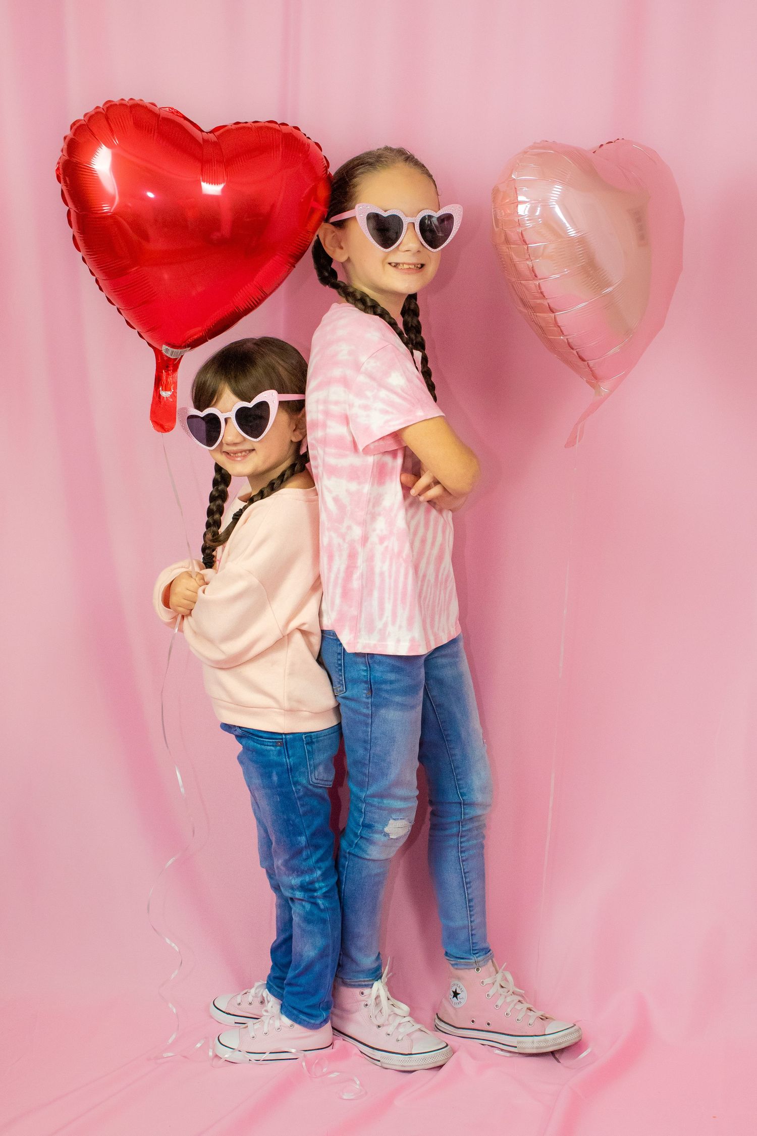 Two children in heart sunglasses stand back-to-back with red and pink heart balloons.