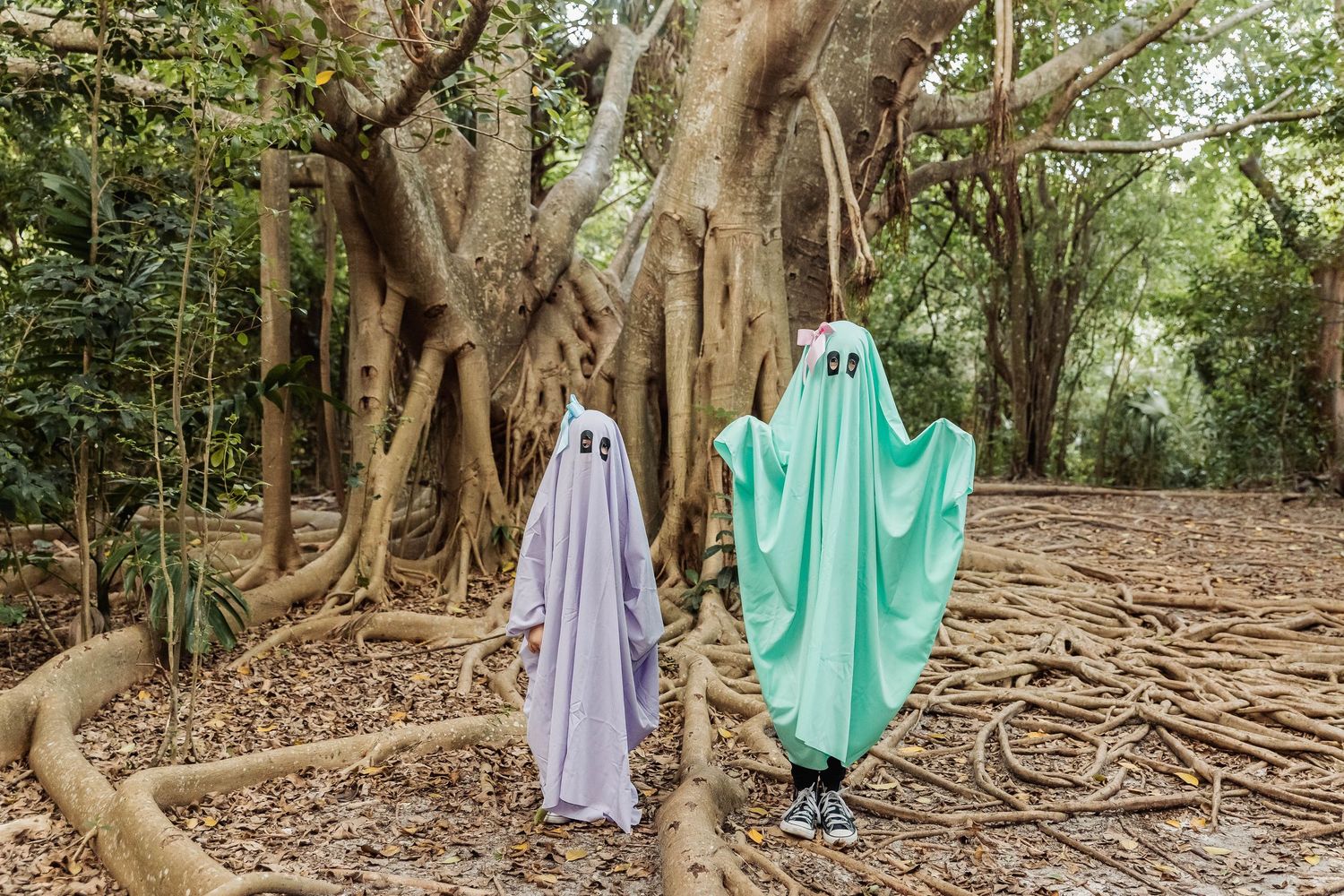 Two ghostly figures in flowing fabric costumes stand among twisted tree roots in a forest setting.