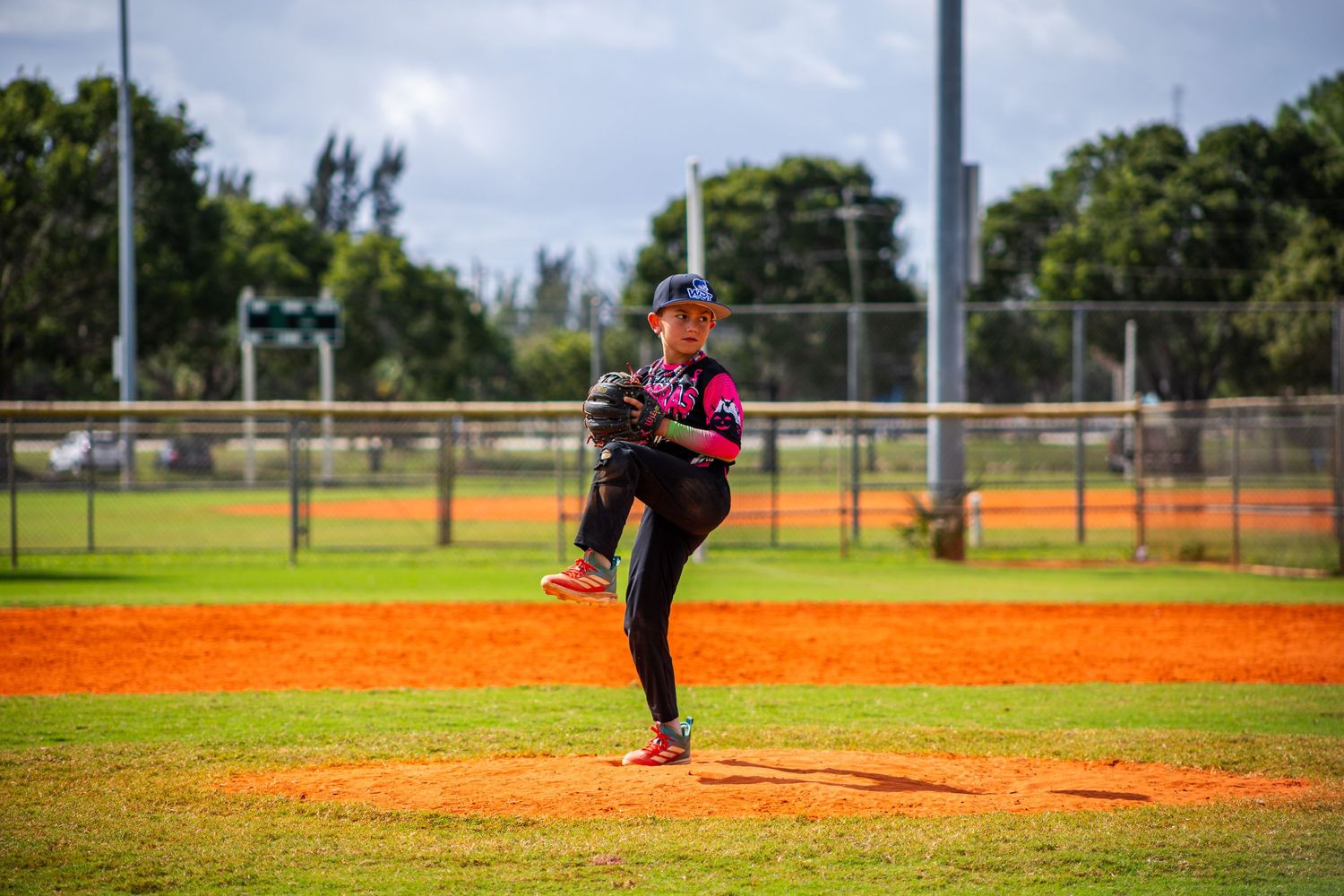 Baseball pitcher in pink jersey winds up for a throw on a sunny day at a baseball field.