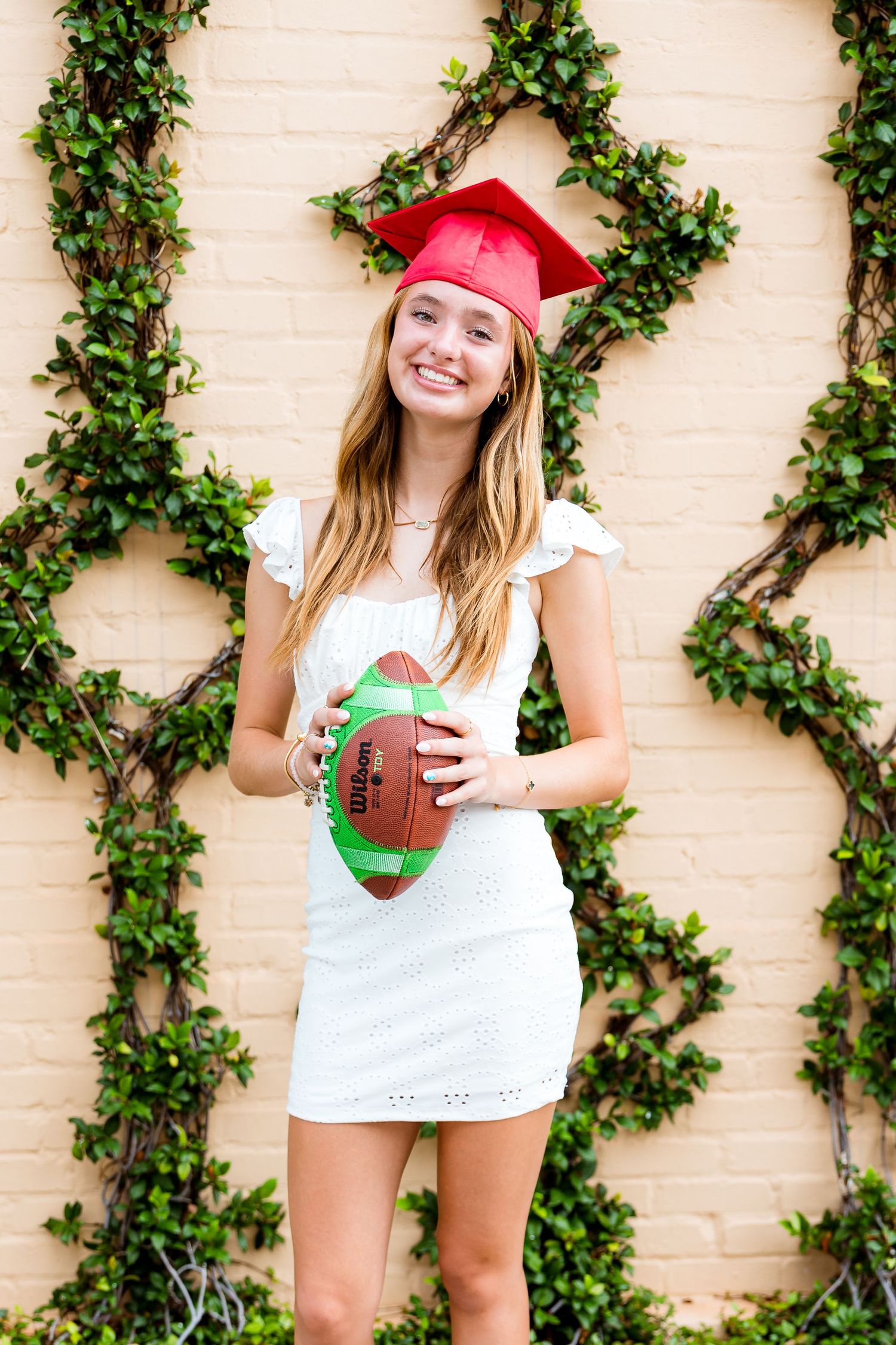 Graduate in red cap and white dress holds football in front of ivy-covered wall during graduation photo shoot.