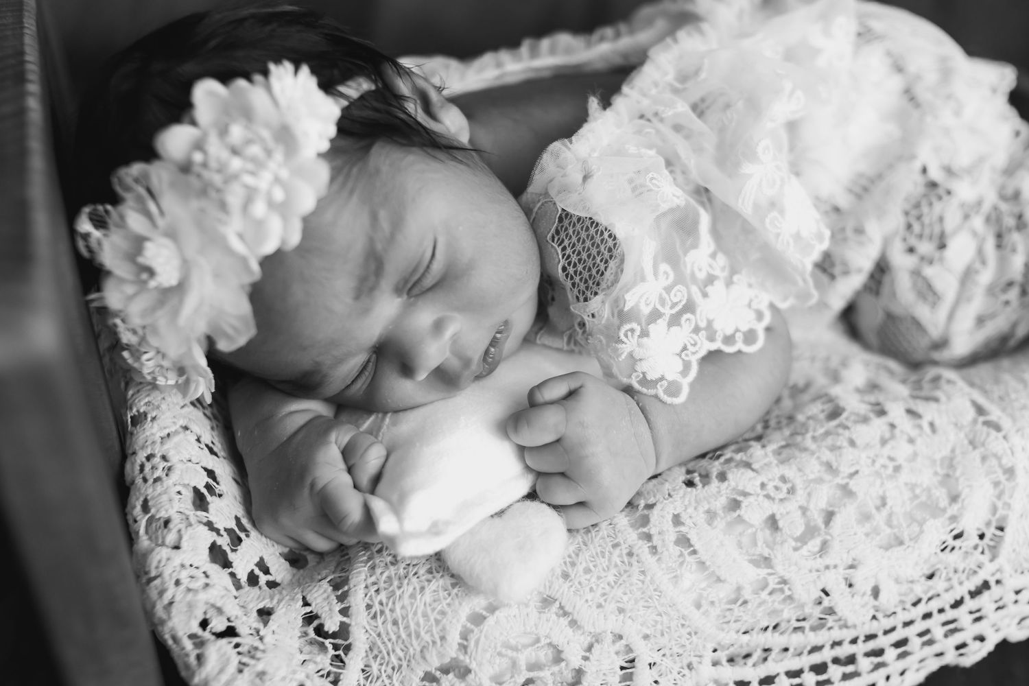 Black and white photo of sleeping newborn in lace outfit and white floral headband on crochet blanket.