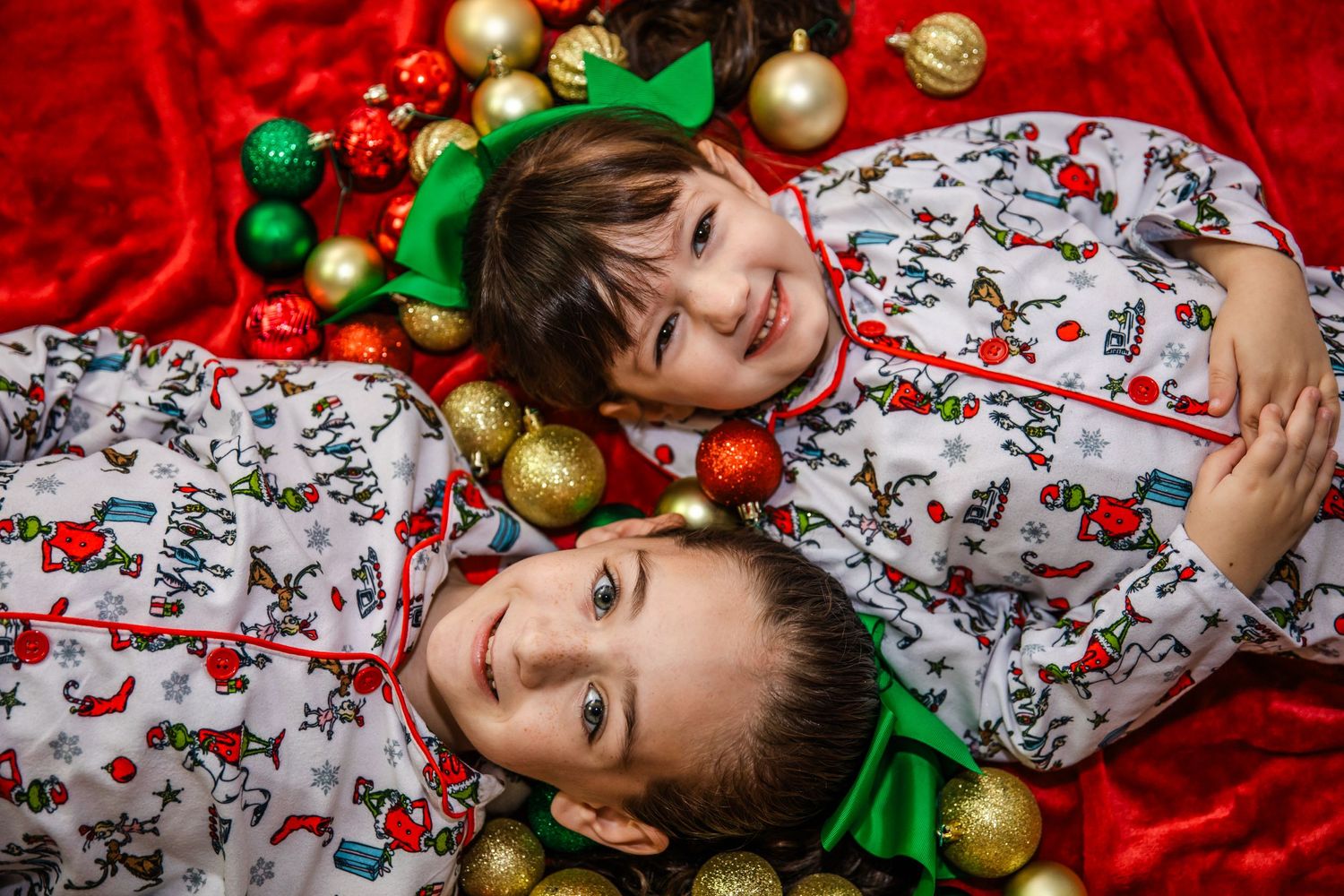 Two children in matching winter pajamas lie on red fabric surrounded by gold and green Christmas ornaments.