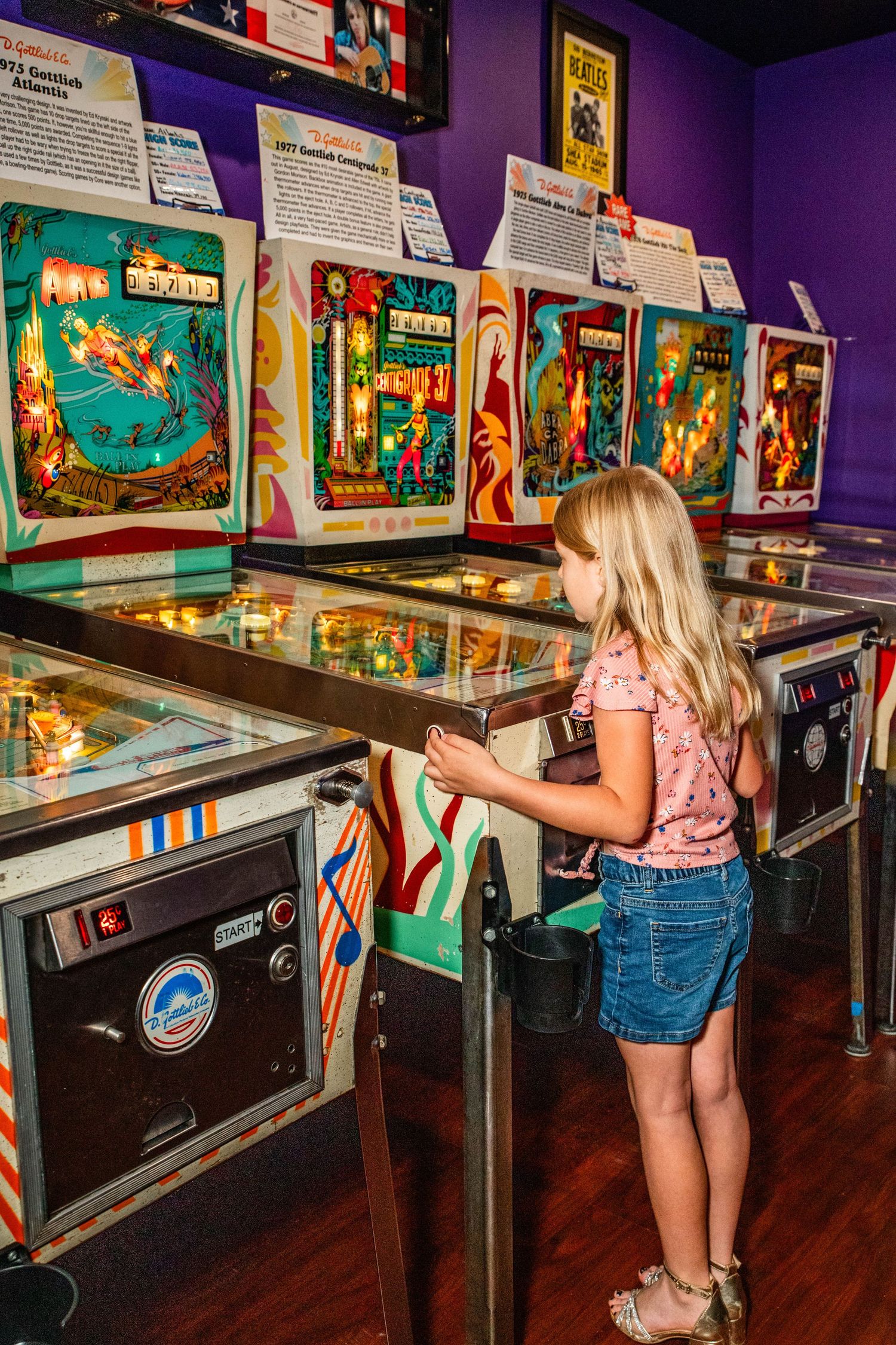 Young girl playing vintage pinball machines at a retro arcade museum with colorful displays.