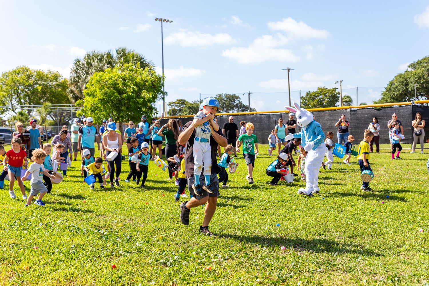 Children and families participate in an outdoor Easter egg hunt on a green field with an Easter Bunny costume character.