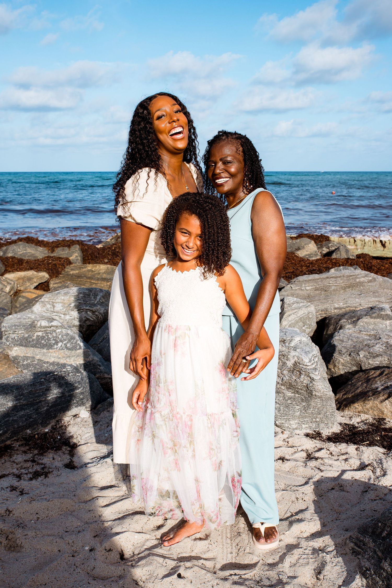 Three people share joyful moments together on a rocky beach with ocean waves and blue skies in the background.