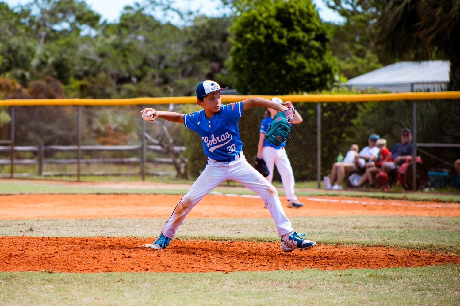 Baseball pitcher in blue jersey mid-delivery on dirt mound during youth baseball game.