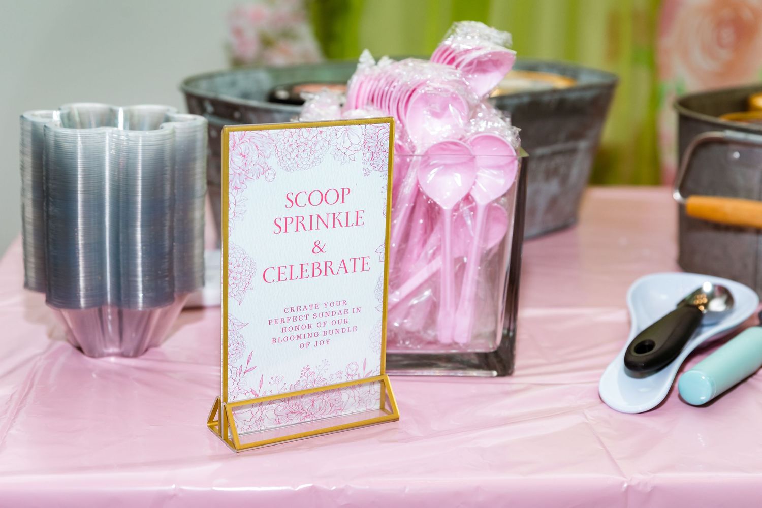 Elegant party decor featuring a gold-framed 'Scoop Sprinkle Celebrate' sign with pink and gray party accessories.