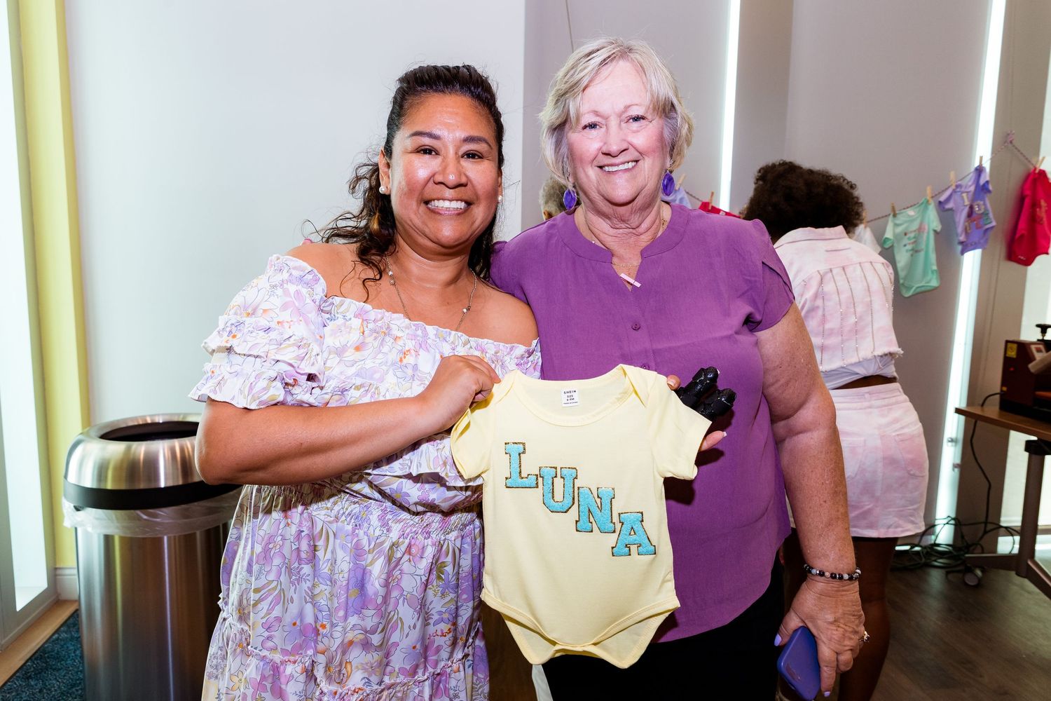 Two attendees display a yellow baby onesie at a shower celebration.
