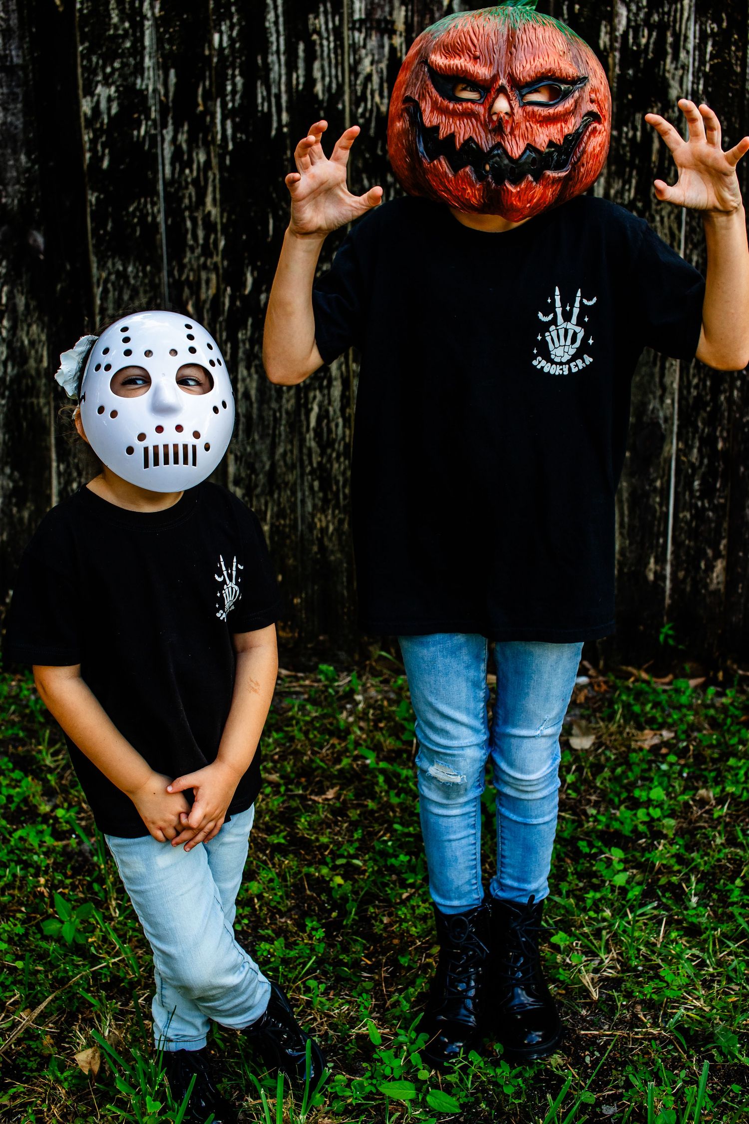 Two children in Halloween masks and matching shirts pose against wooden wall.
