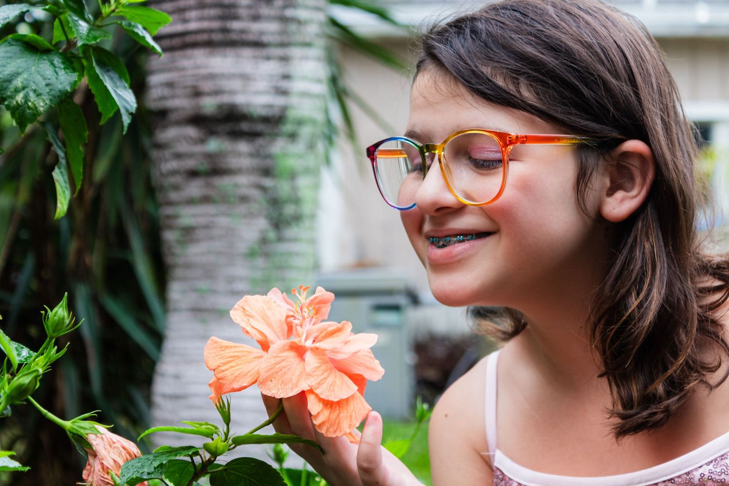 Person wearing orange glasses admires coral flower in garden with palm trees.
