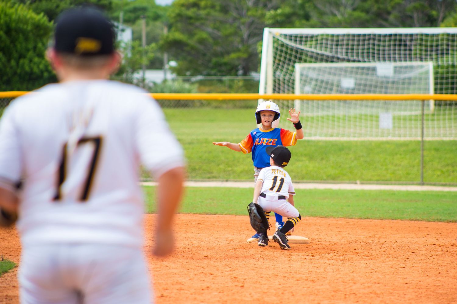 Youth baseball players in action on a dirt infield with a soccer goal visible in the background.