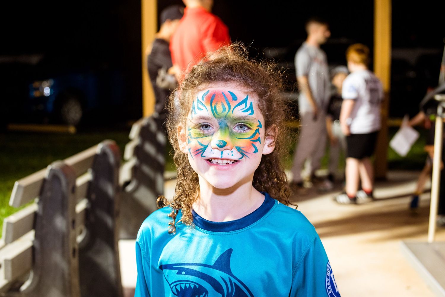 Young person with colorful face paint smiles brightly wearing a blue t-shirt at an outdoor event.