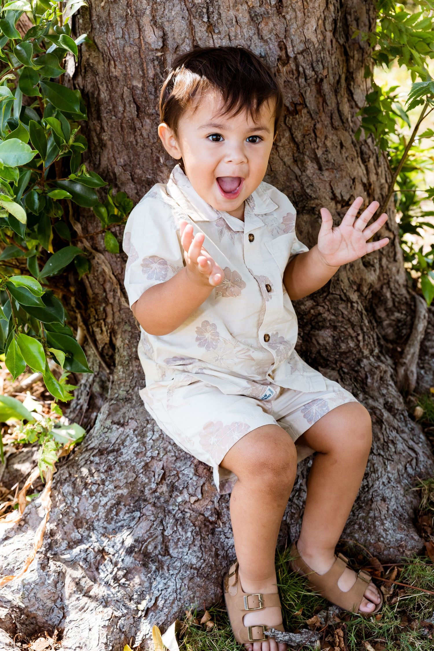 Small child in white outfit sitting against large tree trunk with playful expression and raised hands.