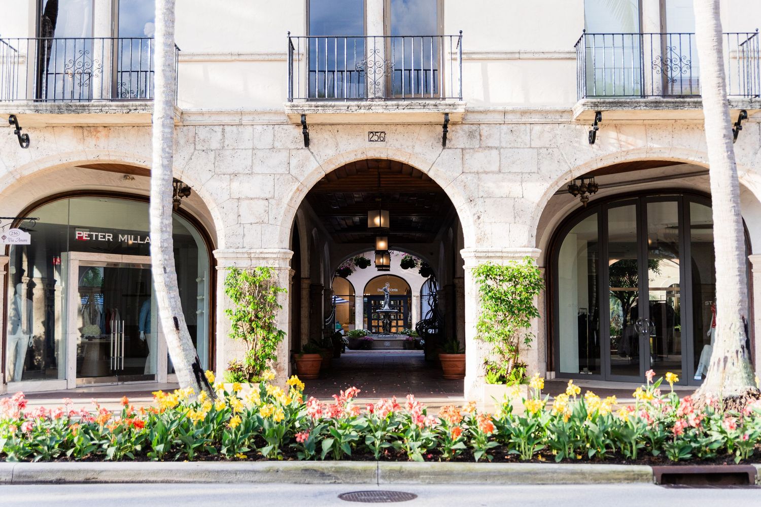Mediterranean style shopping arcade with arched passageway and colorful flower garden along a retail storefront.