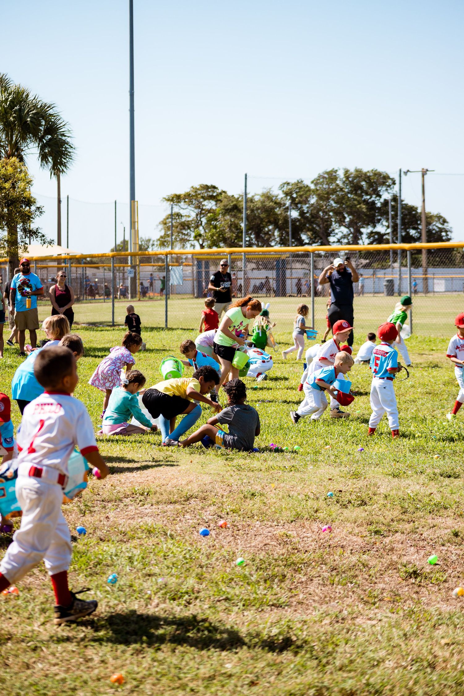 Young children in colorful uniforms run across a grassy field collecting Easter eggs during an outdoor hunt with palm trees visible.