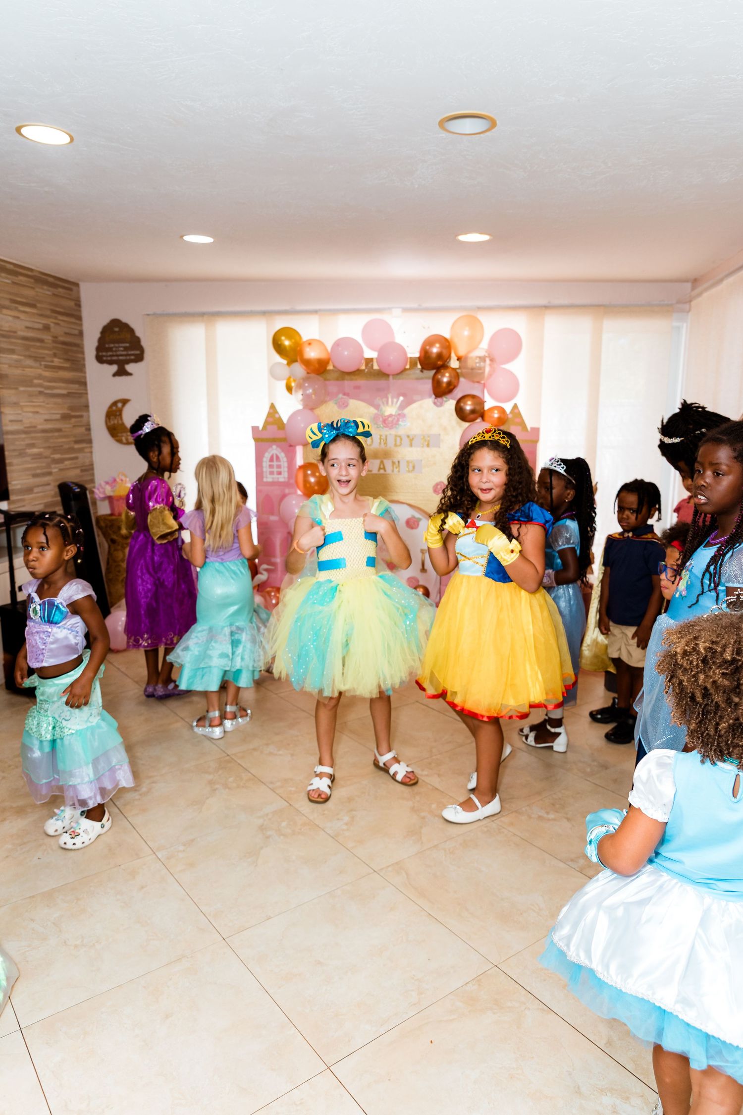 Children in colorful princess costumes dancing at a birthday party with balloon decorations.