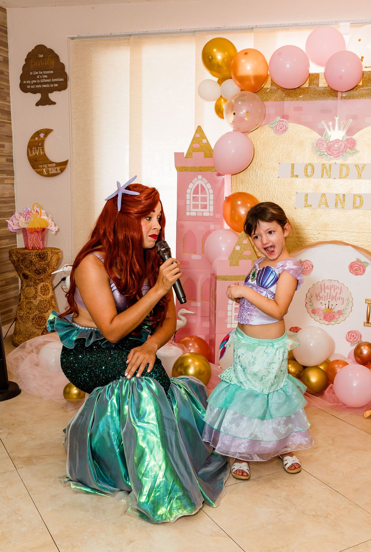 Two people in teal and aqua costumes dance and perform at a magical princess party with pink and gold balloons.