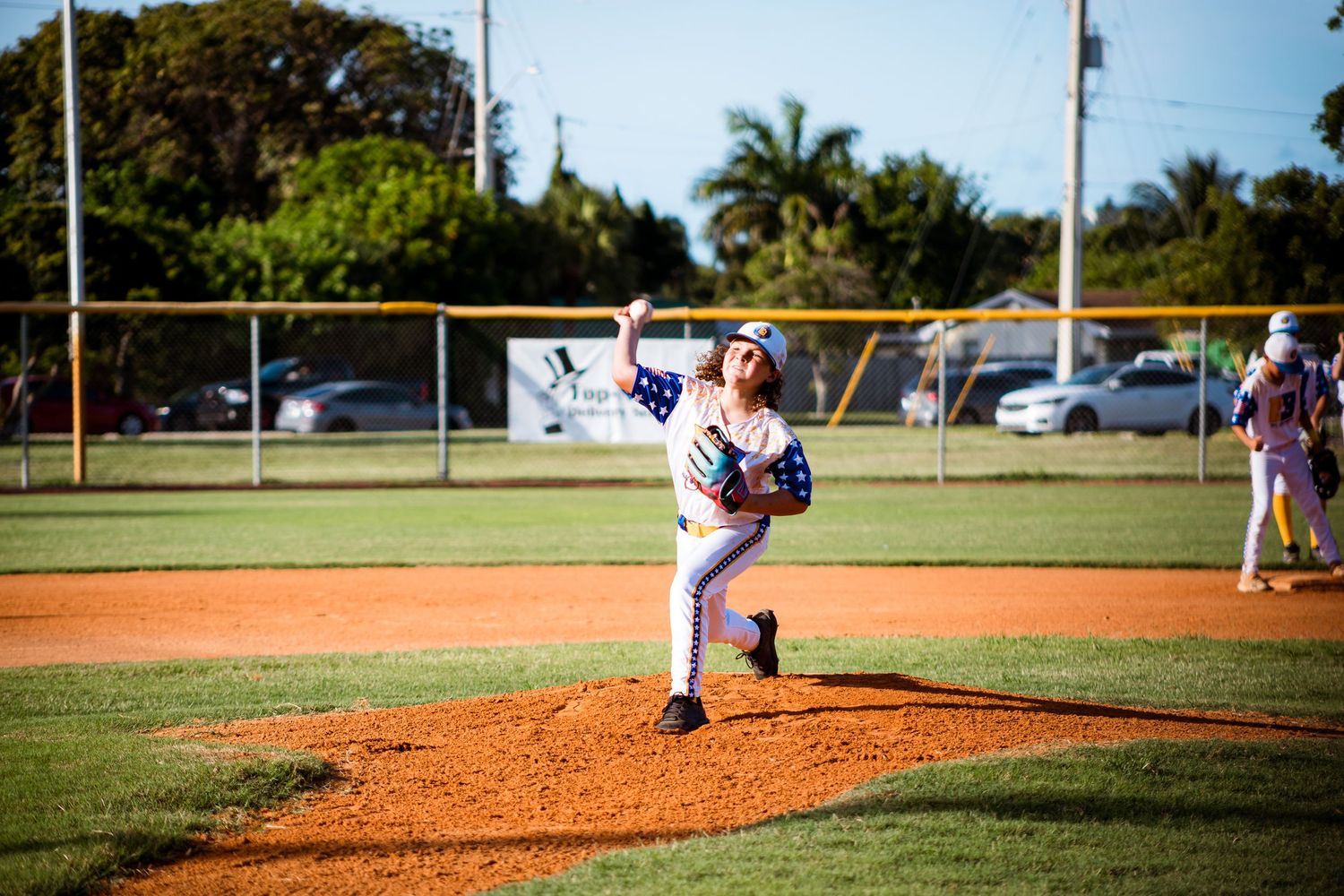 Baseball pitcher in white uniform delivers pitch from mound during daytime game on well-maintained field.