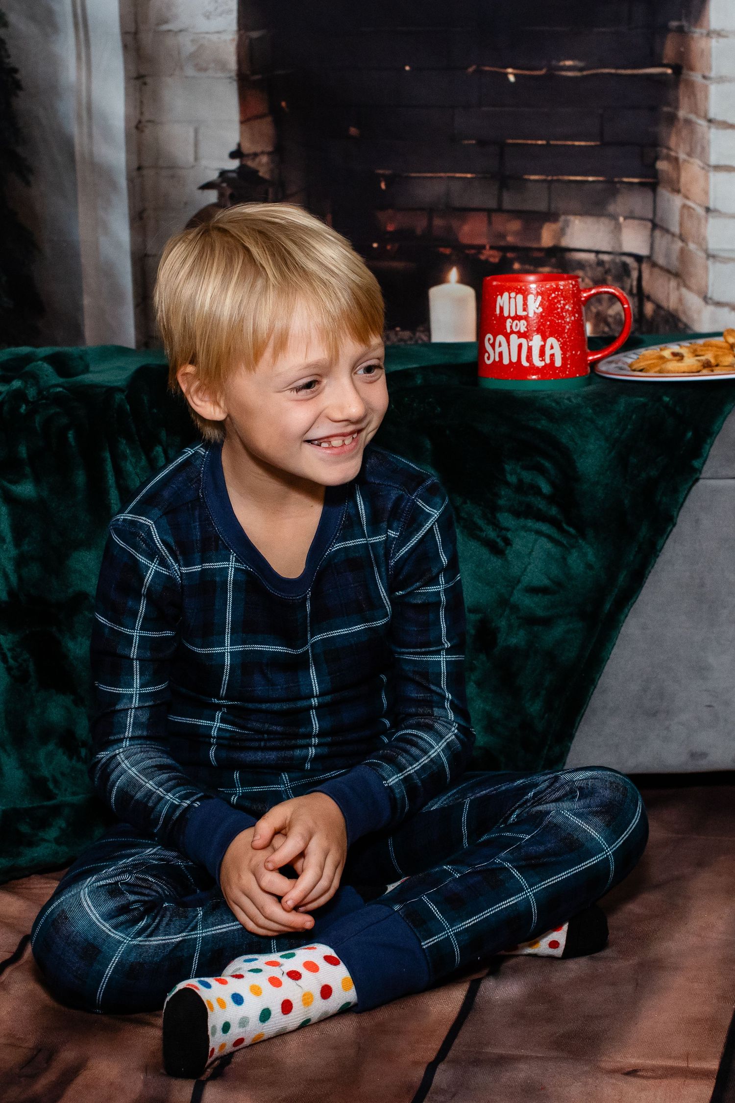 Child in checkered pajamas sits by fireplace with Santa mug and holiday treats.