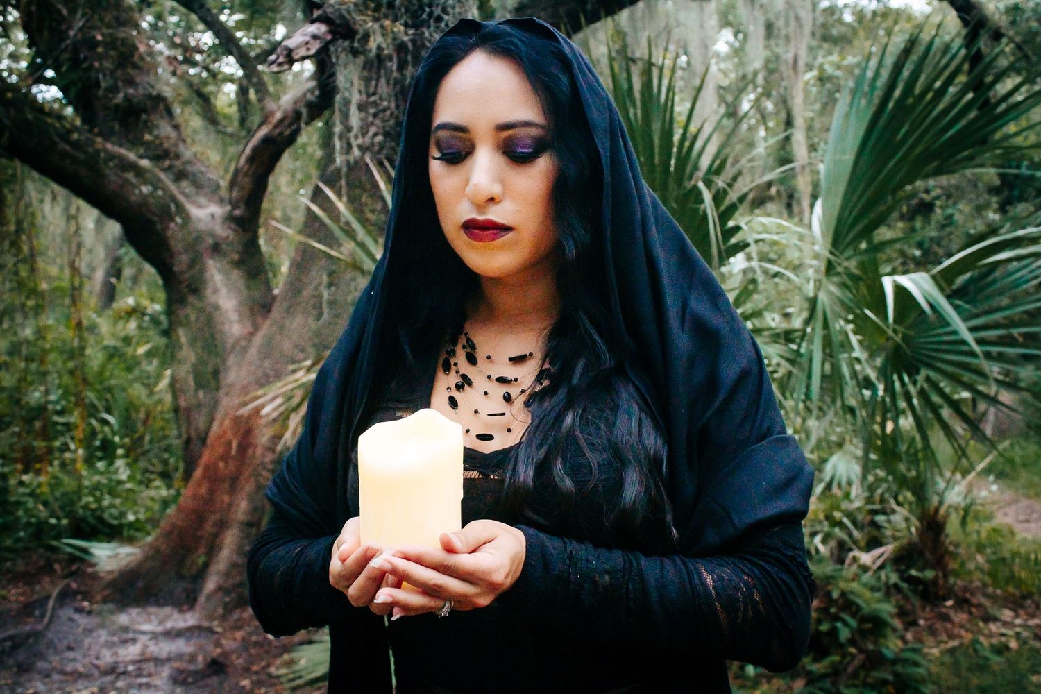 A gothic figure in black holds a white candle against a backdrop of palm fronds and Spanish moss.