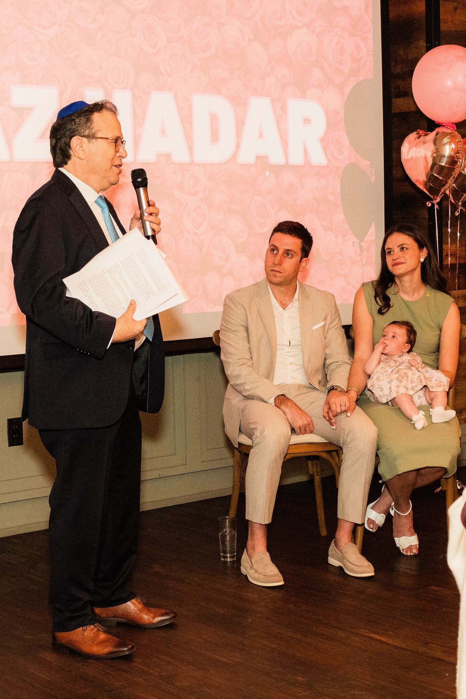 A speaker presenting at an event while others listen seated on stage with pink balloons in the background.