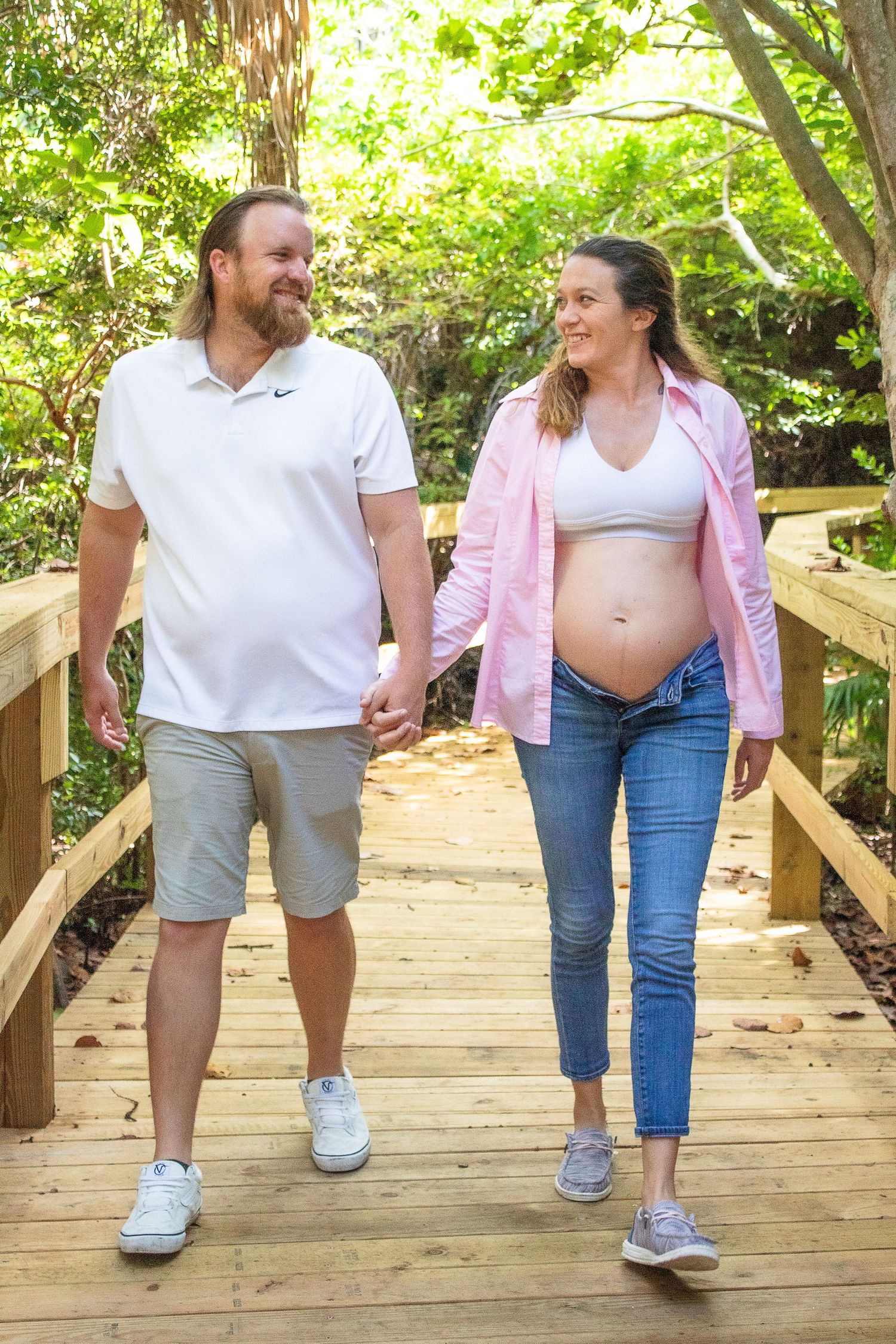 Expecting couple walking hand in hand on a wooden bridge path surrounded by greenery.